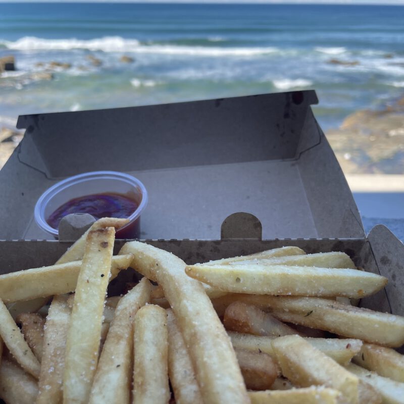 A delicious box of chippies and tomato sauce being held in front of the ocean.